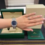 Wrist wearing a silver Rolex Datejust with a blue dial and diamond bezel, green Rolex box in the background.