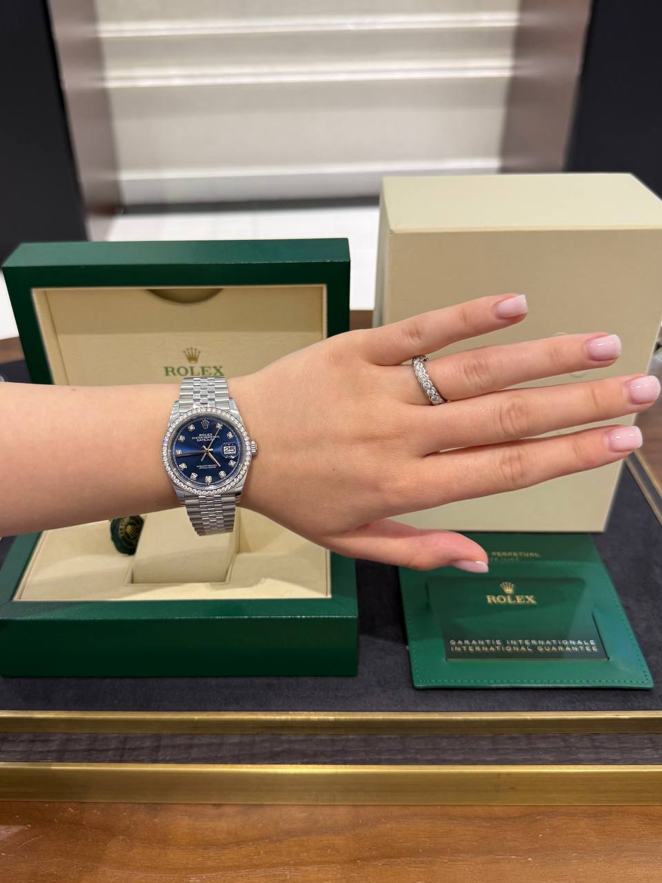Wrist wearing a silver Rolex Datejust with a blue dial and diamond bezel, green Rolex box in the background.
