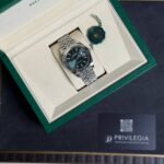 Rolex wristwatch in a beige interior box with a stainless steel bracelet and a green seal tag beside it.