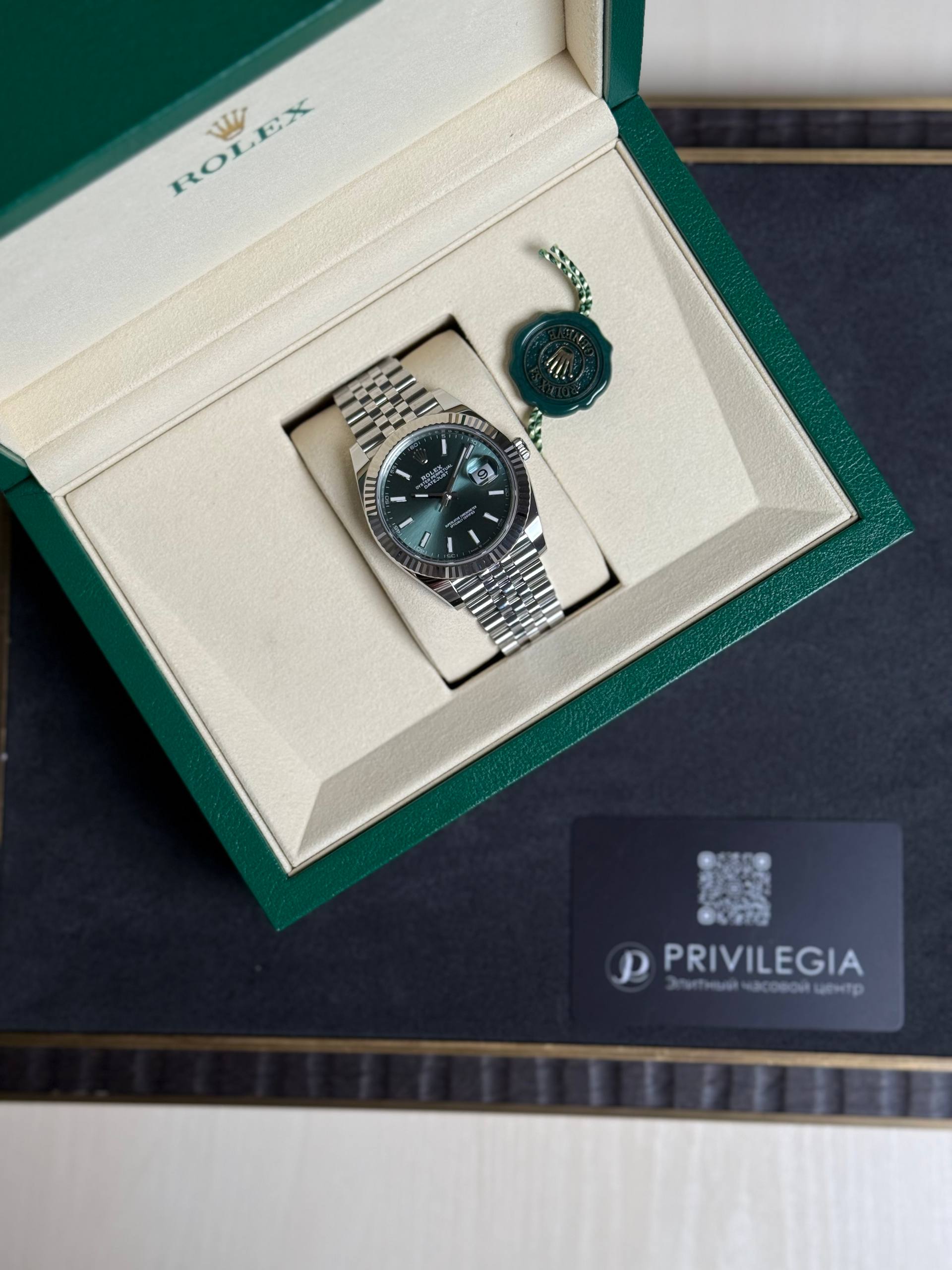 Rolex wristwatch in a beige interior box with a stainless steel bracelet and a green seal tag beside it.