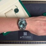 Silver Rolex watch with a green dial on a wrist, beside an open green Rolex box and hanging green seal.