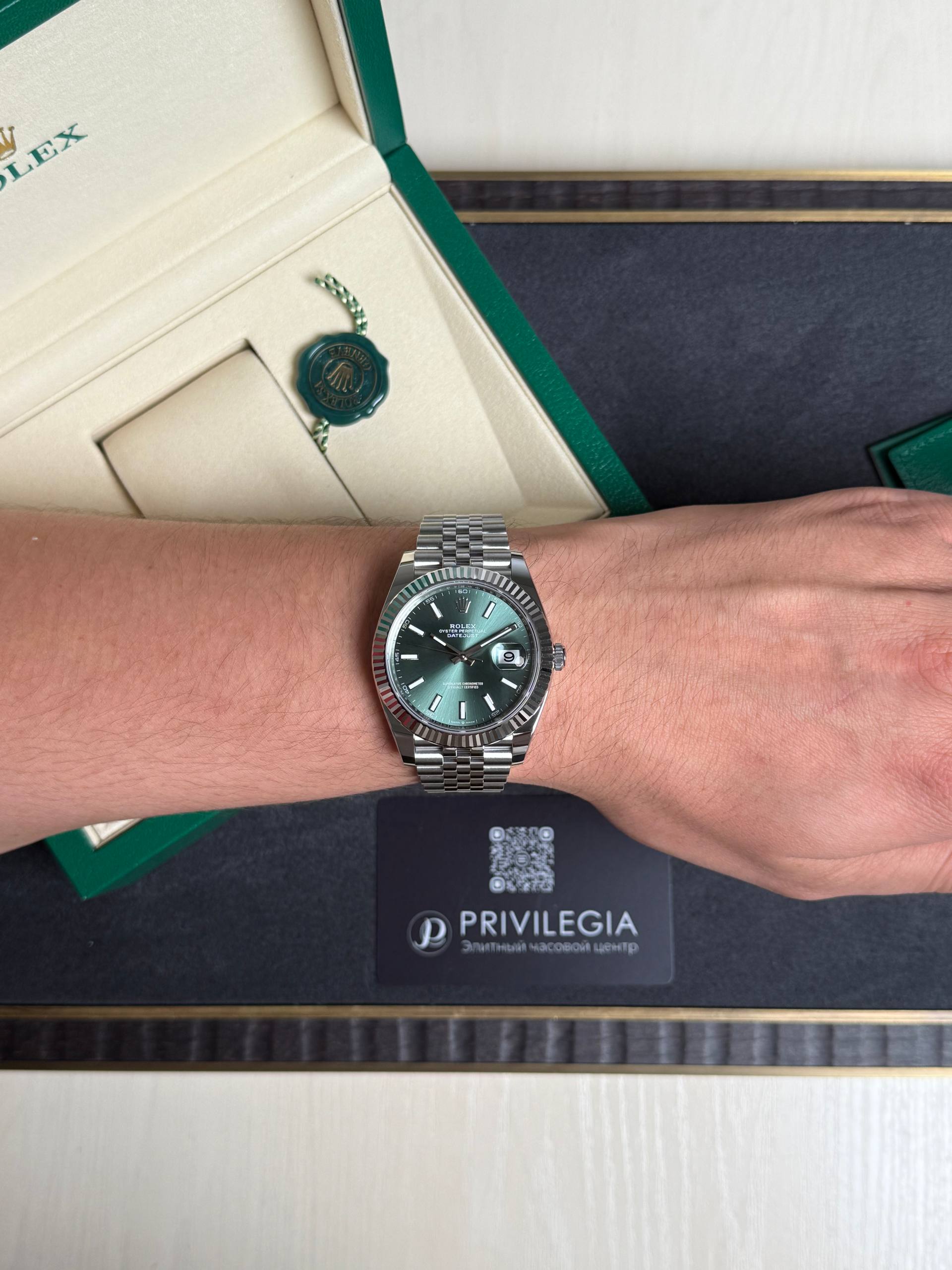 Silver Rolex watch with a green dial on a wrist, beside an open green Rolex box and hanging green seal.