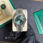 Rolex Oyster Perpetual Datejust stainless steel watch with a green dial and fluted bezel, on a beige cushion inside a green presentation box.