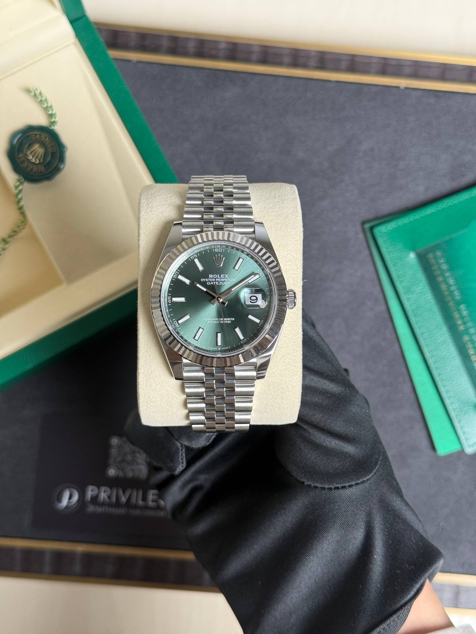 Rolex Oyster Perpetual Datejust stainless steel watch with a green dial and fluted bezel, on a beige cushion inside a green presentation box.