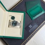 Rolex watch in a green presentation box with a metal bracelet, alongside green authentication cards on a dark surface.