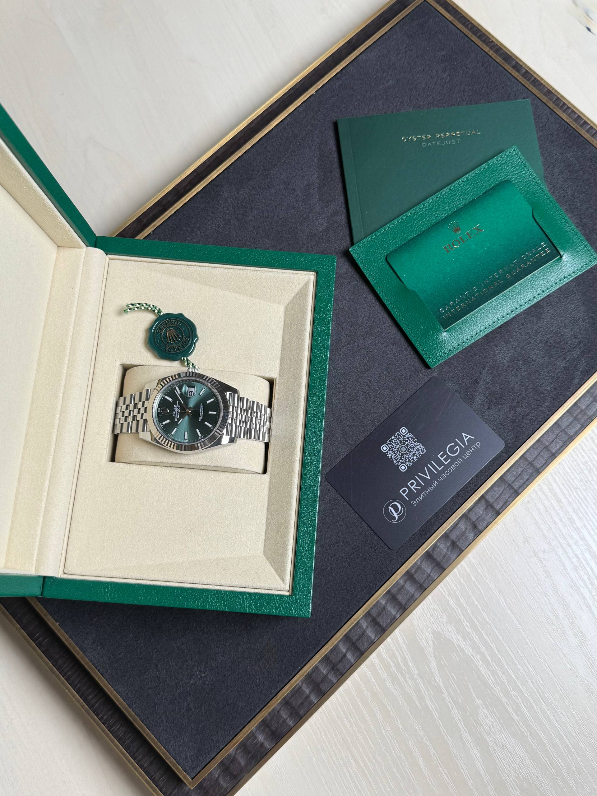 Rolex watch in a green presentation box with a metal bracelet, alongside green authentication cards on a dark surface.