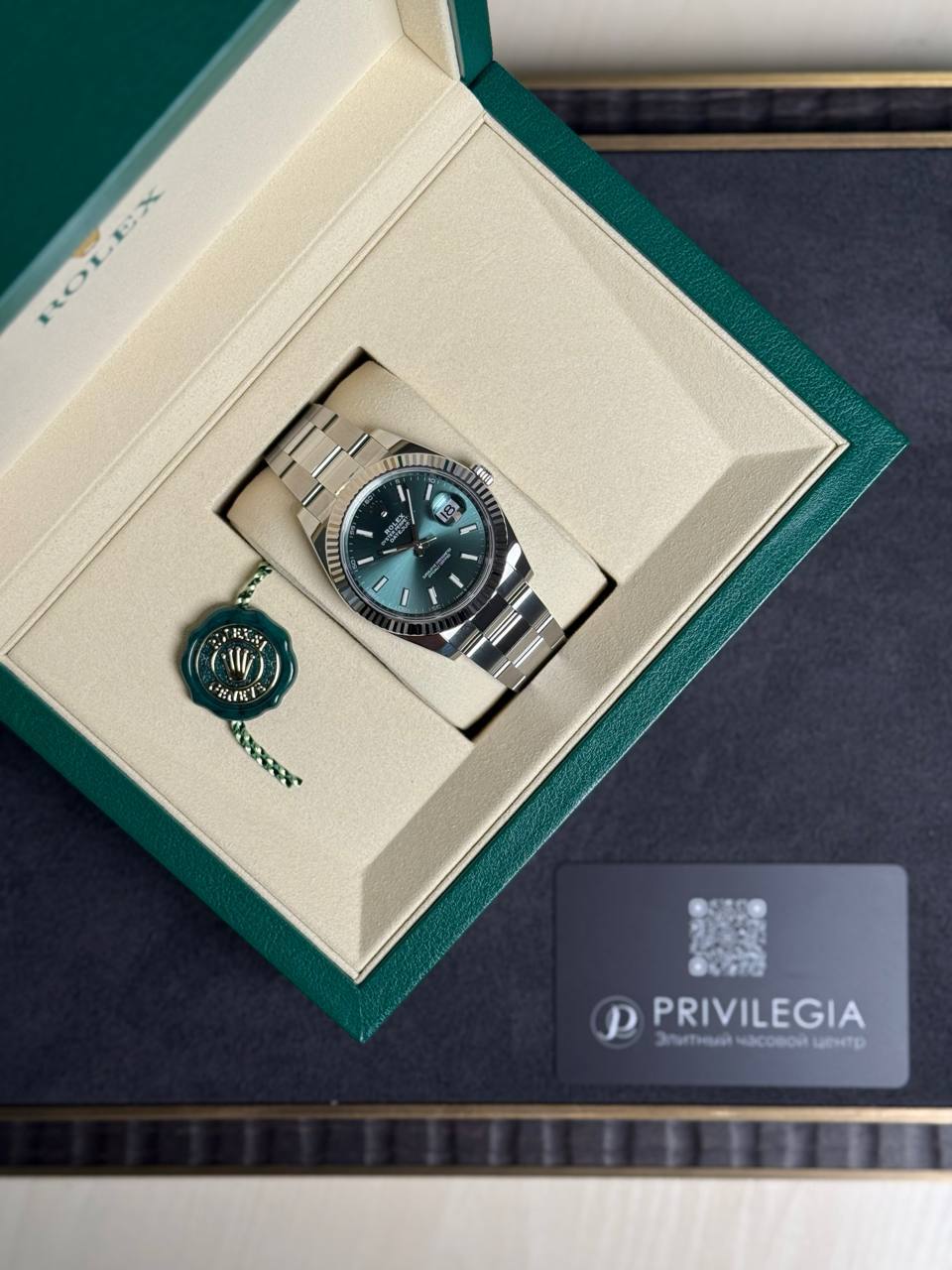 Rolex stainless steel wristwatch with green dial in its beige presentation box, with green seal tag nearby.