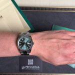 Wrist wearing a stainless steel Rolex Datejust with a green dial and date window, beside a green box and Privilegia card.