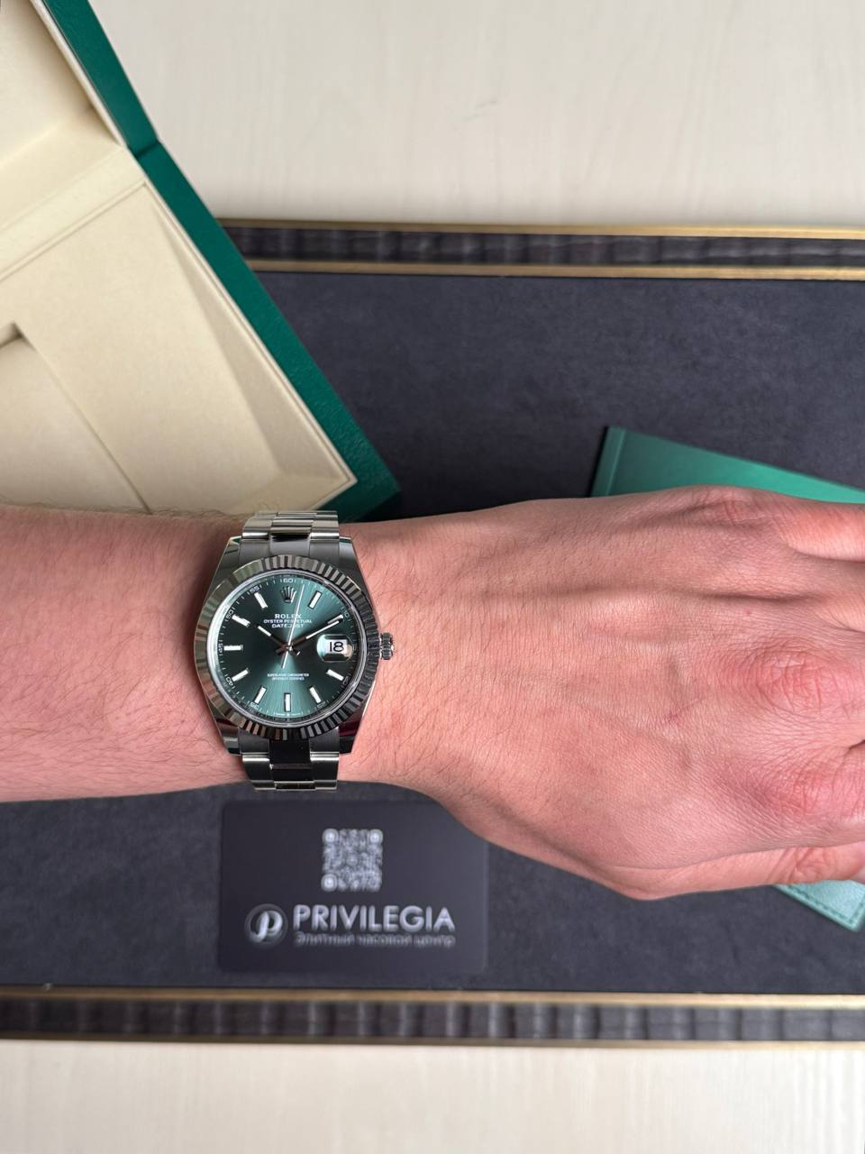 Wrist wearing a stainless steel Rolex Datejust with a green dial and date window, beside a green box and Privilegia card.