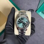 Rolex Datejust wristwatch with a green dial and fluted bezel on a beige display cushion, held by a gloved hand.