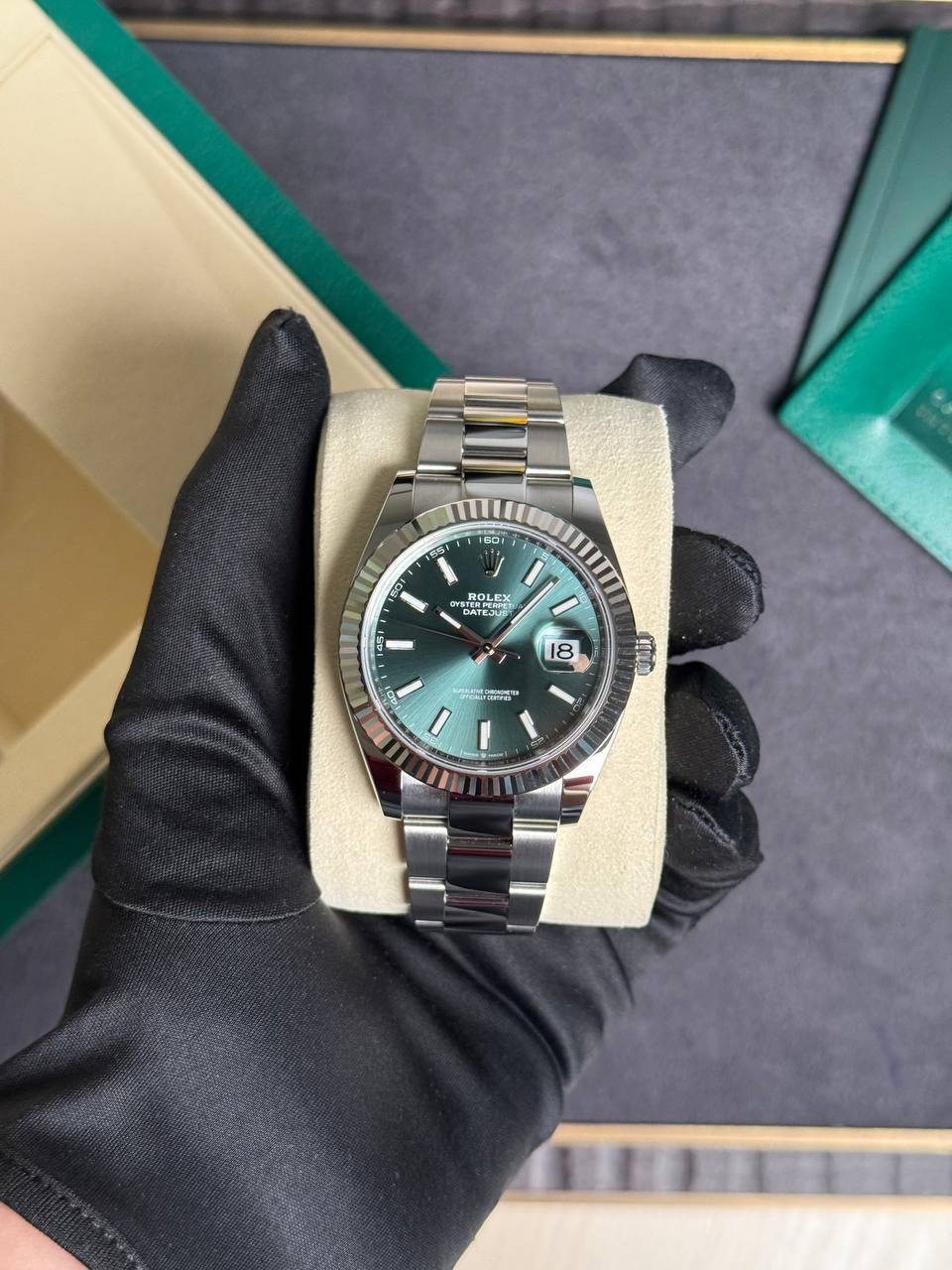 Rolex Datejust wristwatch with a green dial and fluted bezel on a beige display cushion, held by a gloved hand.