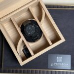 Black Hublot wristwatch resting on a black cushion inside a light wooden display box with compartments.