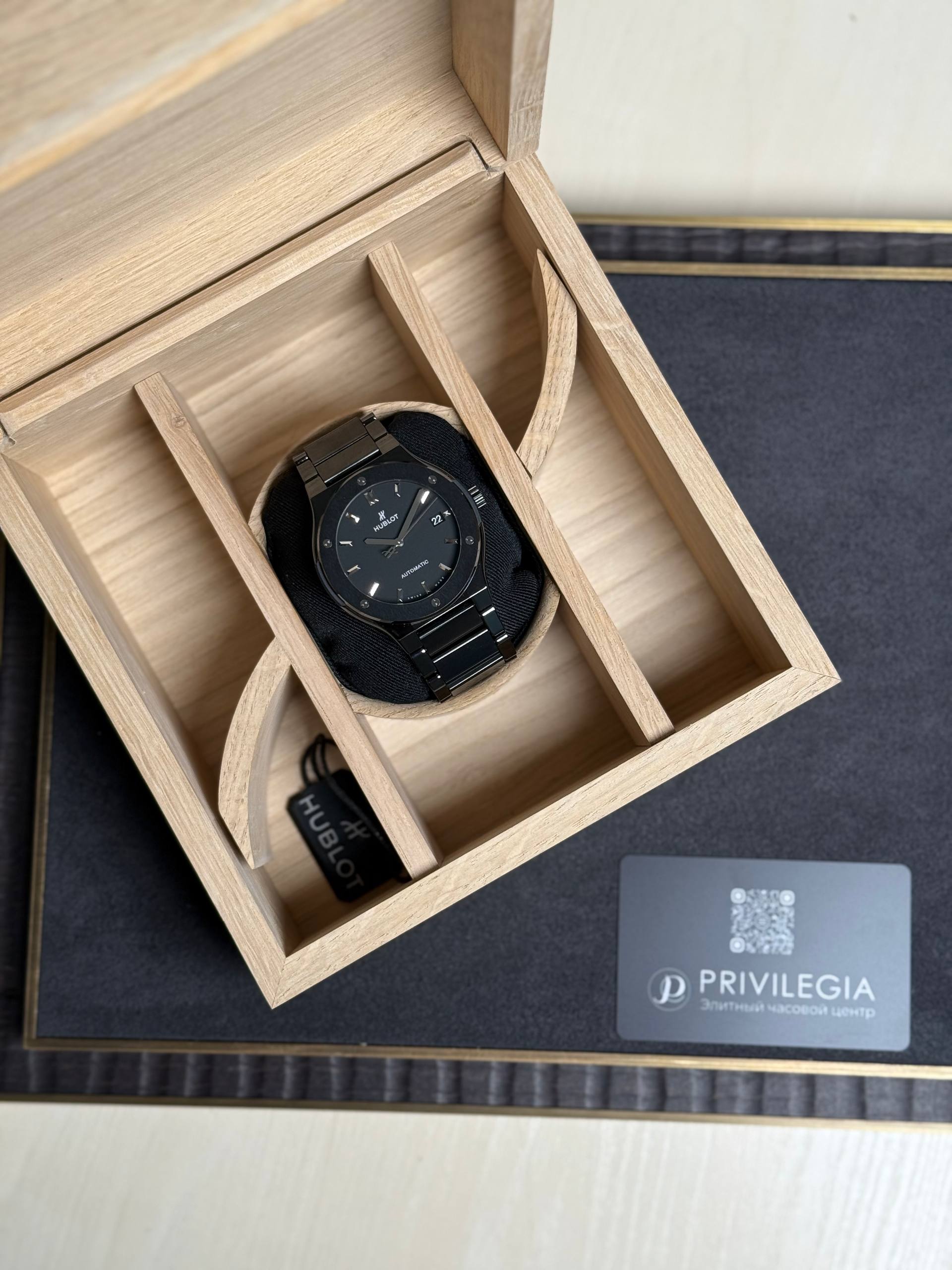 Black Hublot wristwatch resting on a black cushion inside a light wooden display box with compartments.