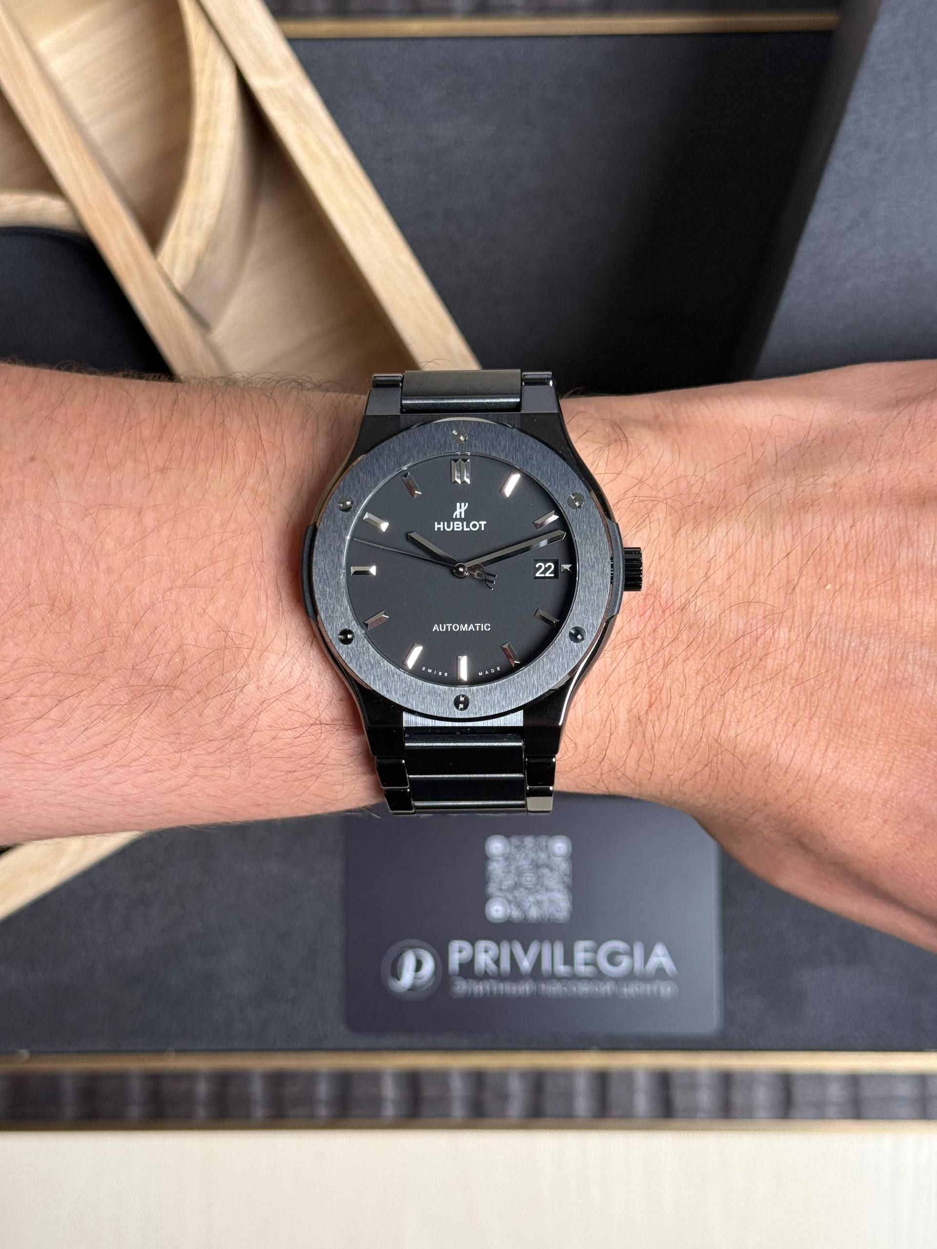 Black Hublot automatic wristwatch on a hairy wrist, date shows 22, with a metal bracelet and dark dial in focus.