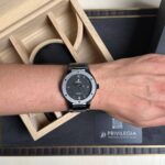 Wrist wearing a black stainless steel Hublot watch with a gray bezel inside a wooden display box interior.