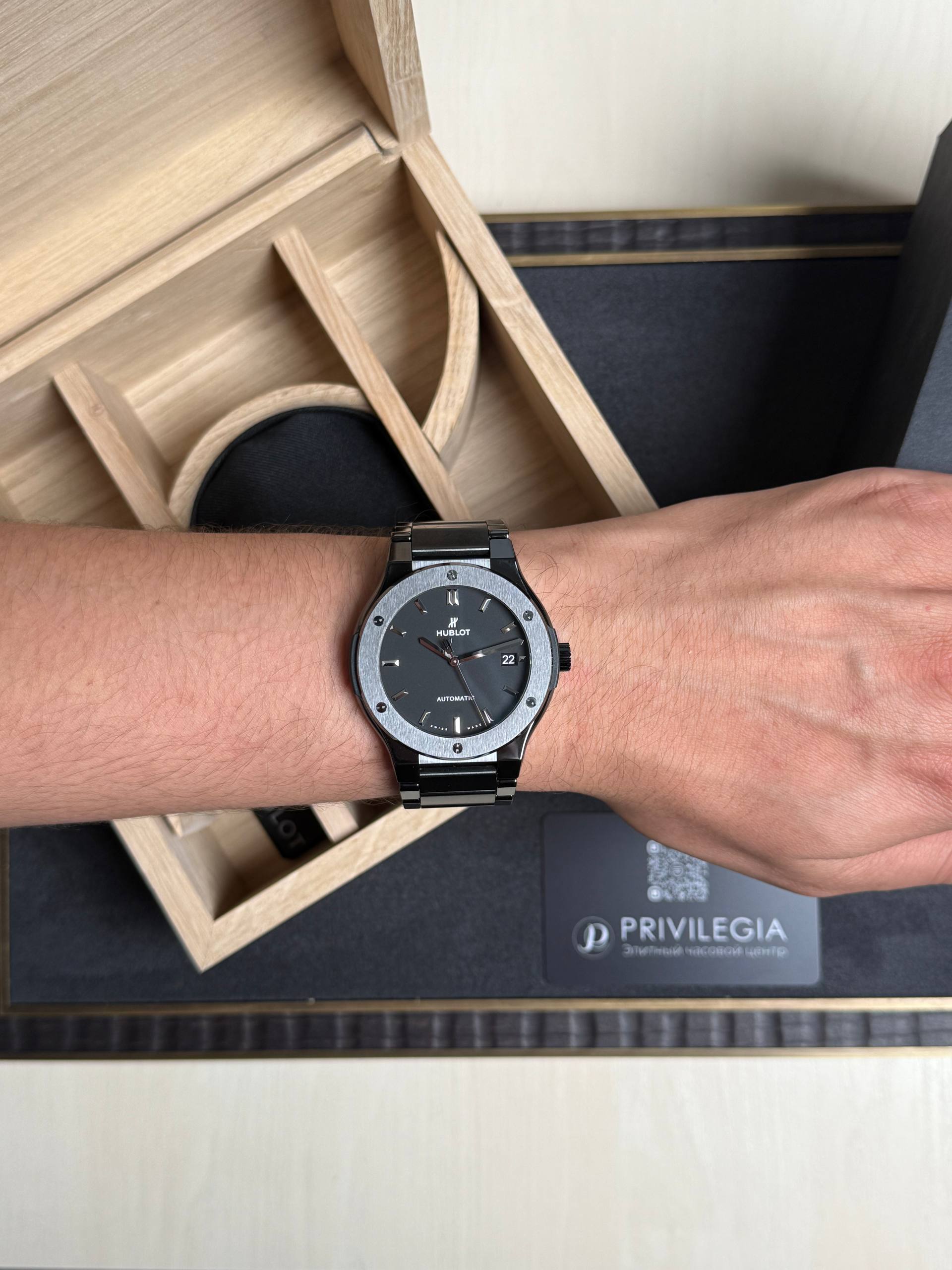 Wrist wearing a black stainless steel Hublot watch with a gray bezel inside a wooden display box interior.