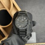 Black metal wristwatch held by gloved hand, Hublot logo visible, in a display box background.
