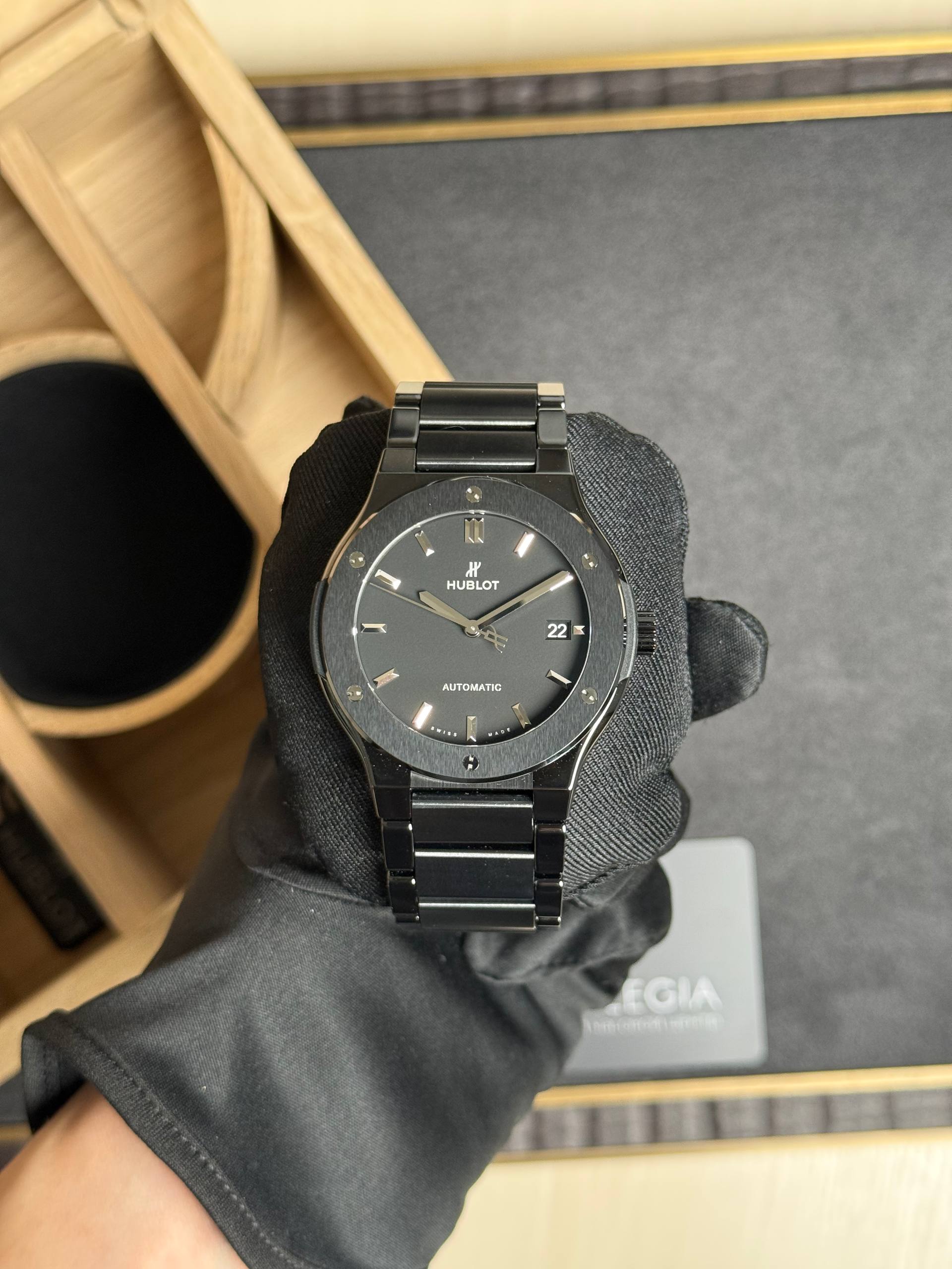 Black metal wristwatch held by gloved hand, Hublot logo visible, in a display box background.