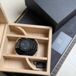 Black Hublot wristwatch in a light wooden display box with a black strap and tag nearby.