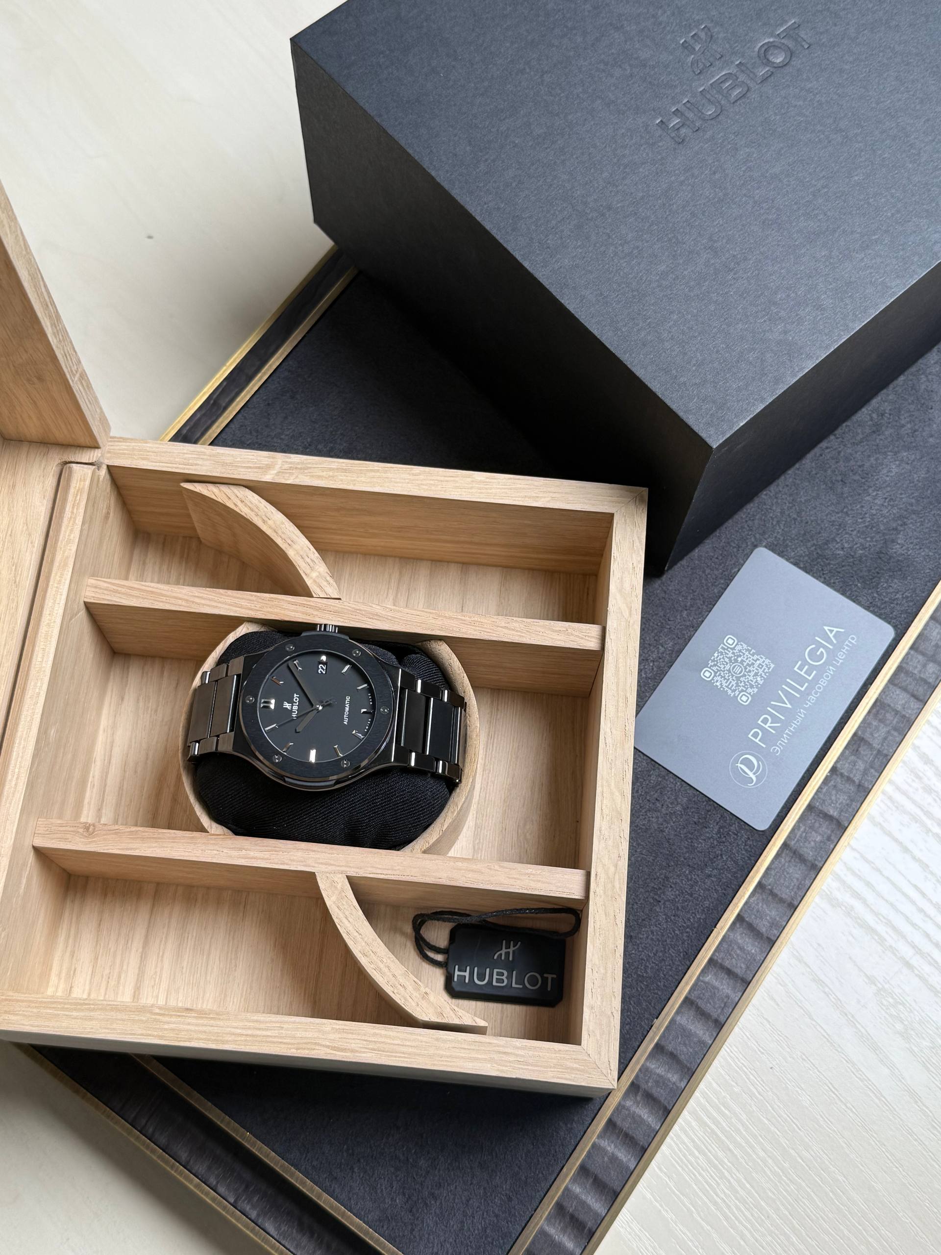 Black Hublot wristwatch in a light wooden display box with a black strap and tag nearby.