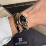 Person's wrist wearing a black Bulgari watch with diamond-studded bezel and a metallic bracelet, against a dark Bulgari box.