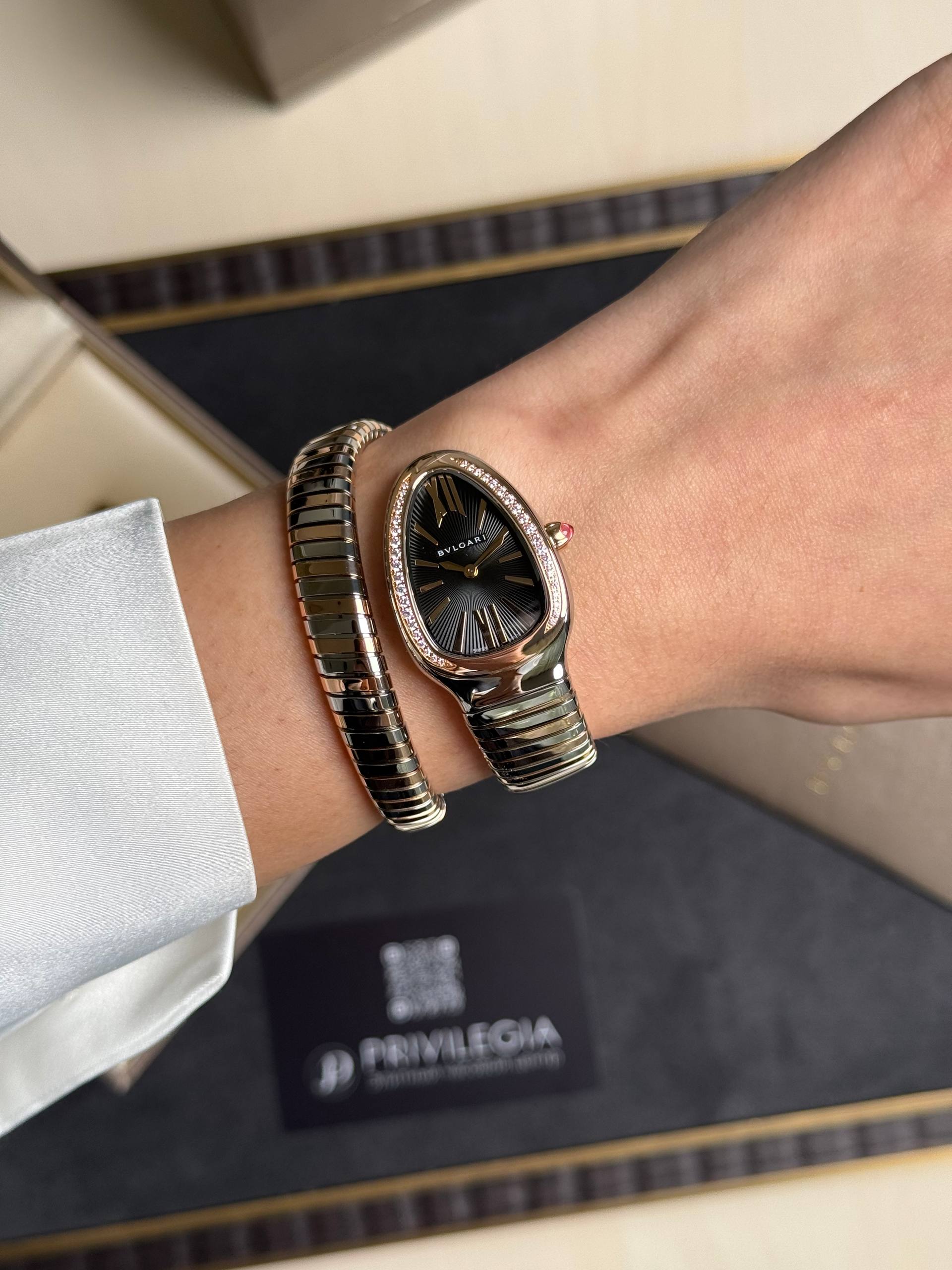 Person's wrist wearing a black Bulgari watch with diamond-studded bezel and a metallic bracelet, against a dark Bulgari box.