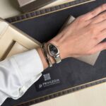 Wrist wearing a silver-toned wristwatch with a metal bracelet, resting on a black display tray inside a gift box.