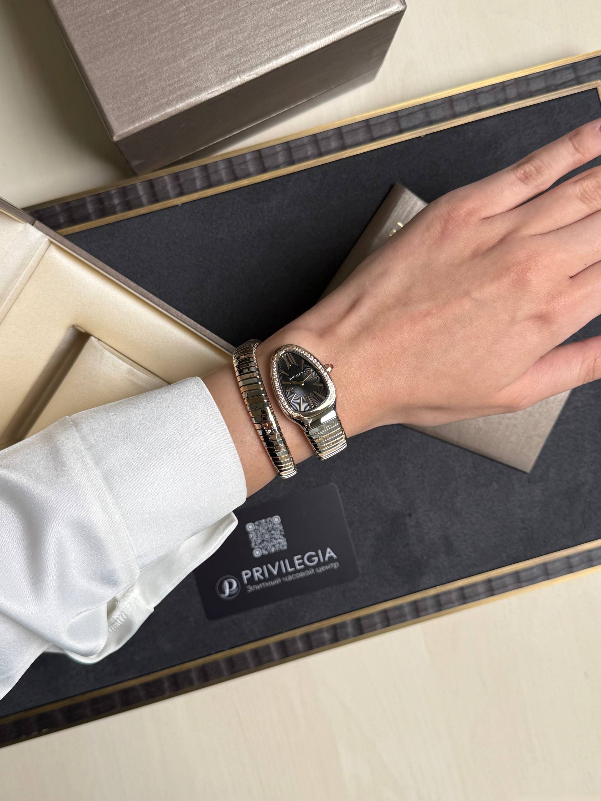 Wrist wearing a silver-toned wristwatch with a metal bracelet, resting on a black display tray inside a gift box.