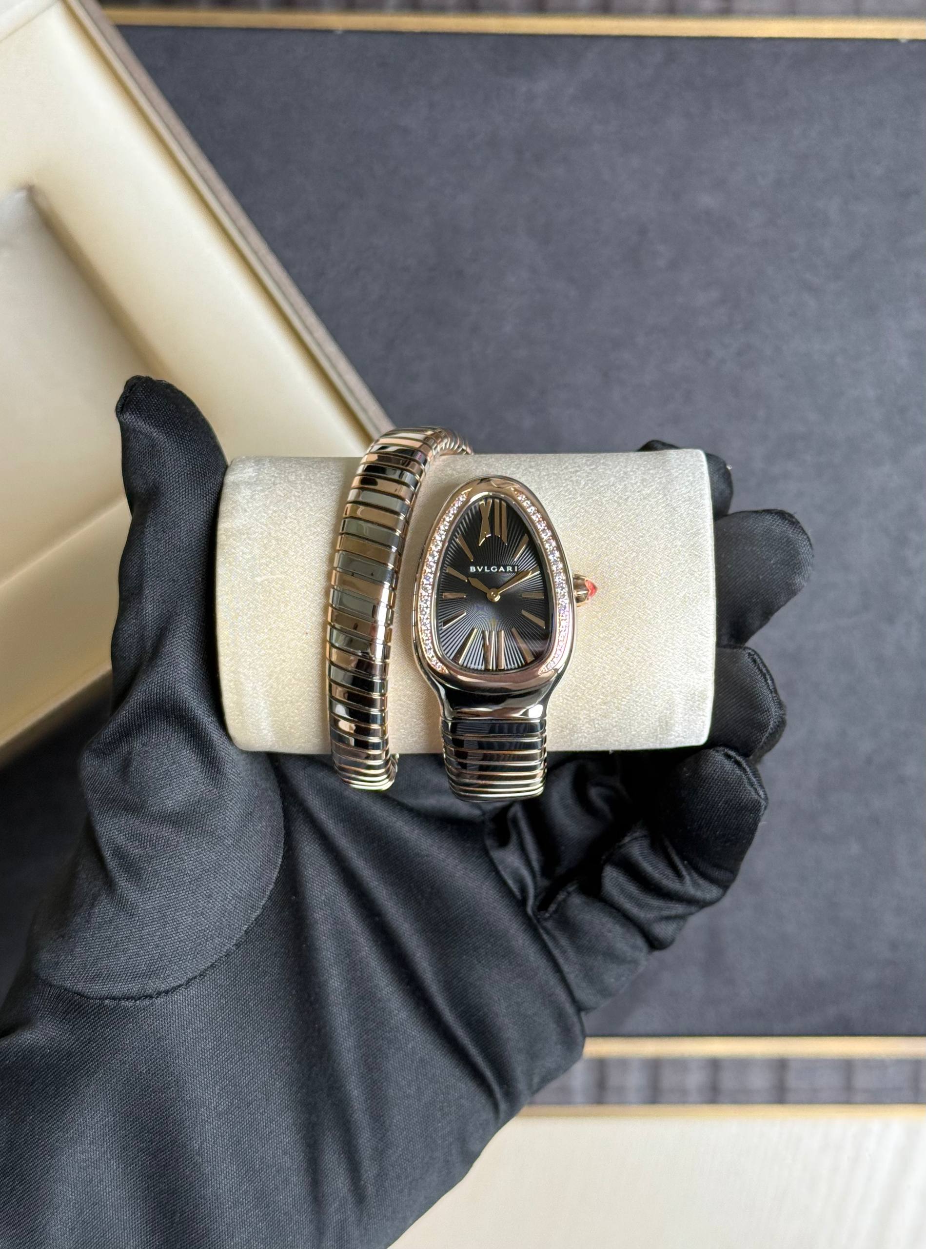 Bulgari wristwatch with an oval black dial and diamond bezel, held in a gloved hand on a beige cushioned case.