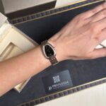 Wristwatch on a woman's left wrist: black dial, diamond bezel, silver bracelet, resting on a jewelry box with a black Privilegia card nearby