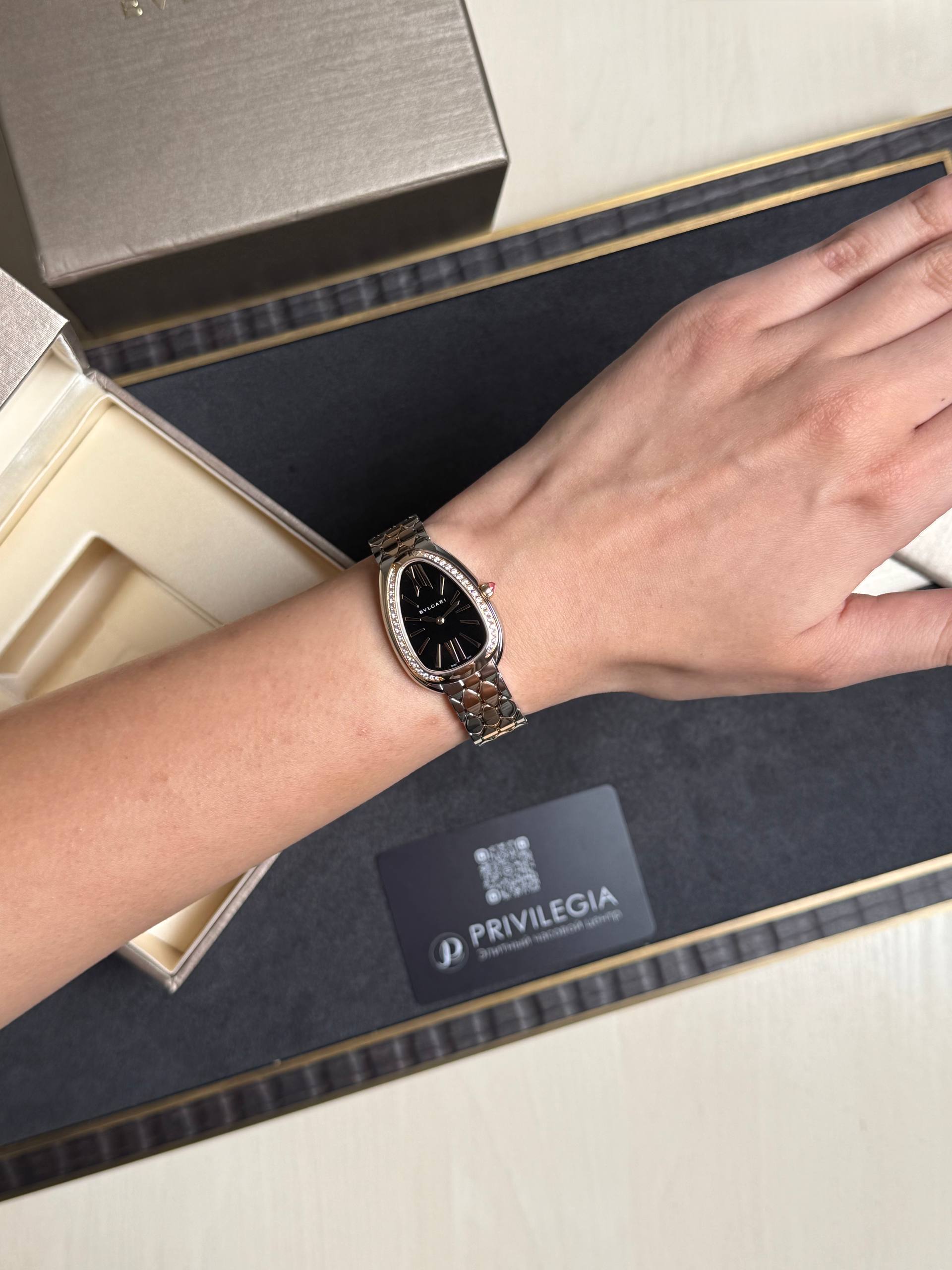 Wristwatch on a woman's left wrist: black dial, diamond bezel, silver bracelet, resting on a jewelry box with a black Privilegia card nearby