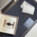 Two-tone BVLGARI wristwatch with a black dial in a cream display box, surrounded by elegant packaging.