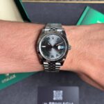 Wrist wearing a silver Rolex Datejust with a fluted bezel and green Roman numerals, inside a Rolex box
