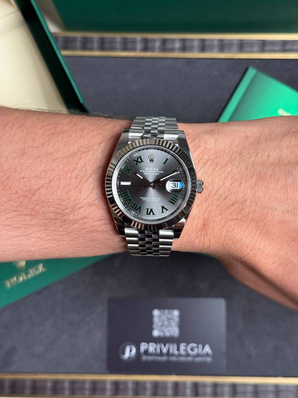 Wrist wearing a silver Rolex Datejust with a fluted bezel and green Roman numerals, inside a Rolex box
