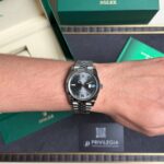 Wrist wearing a silver Rolex Datejust with a dark dial, Roman numerals, and date window, against green Rolex boxes.