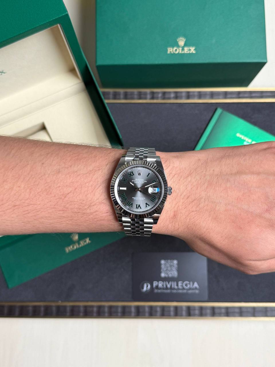 Wrist wearing a silver Rolex Datejust with a dark dial, Roman numerals, and date window, against green Rolex boxes.