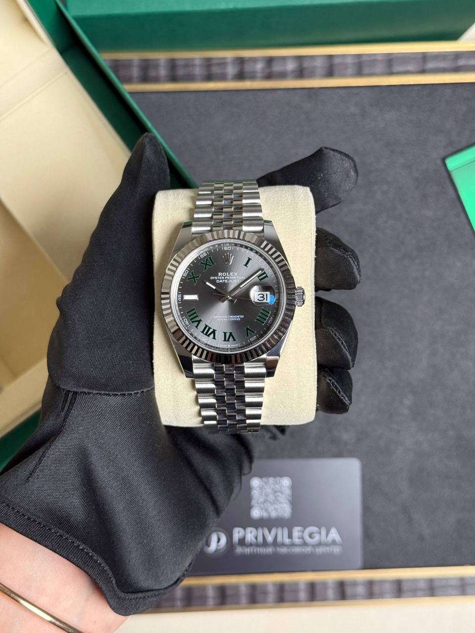 Rolex Datejust on a cushion with a gray dial and green Roman numerals, stainless steel bracelet in a display box.