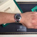 Wrist wearing a stainless steel Rolex watch with a black dial and red/blue bezel, placed on a green presentation box and Privilegia card nearby.