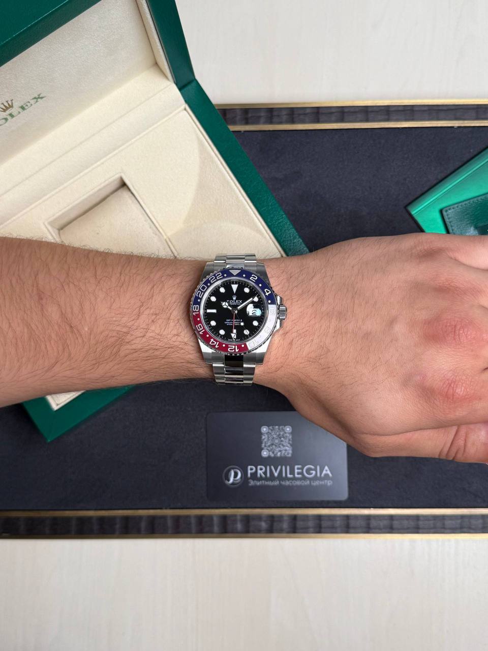 Wrist wearing a stainless steel Rolex watch with a black dial and red/blue bezel, placed on a green presentation box and Privilegia card nearby.