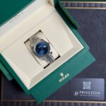 Rolex watch in an open beige interior box with a blue dial and silver bracelet, set inside a green outer box.