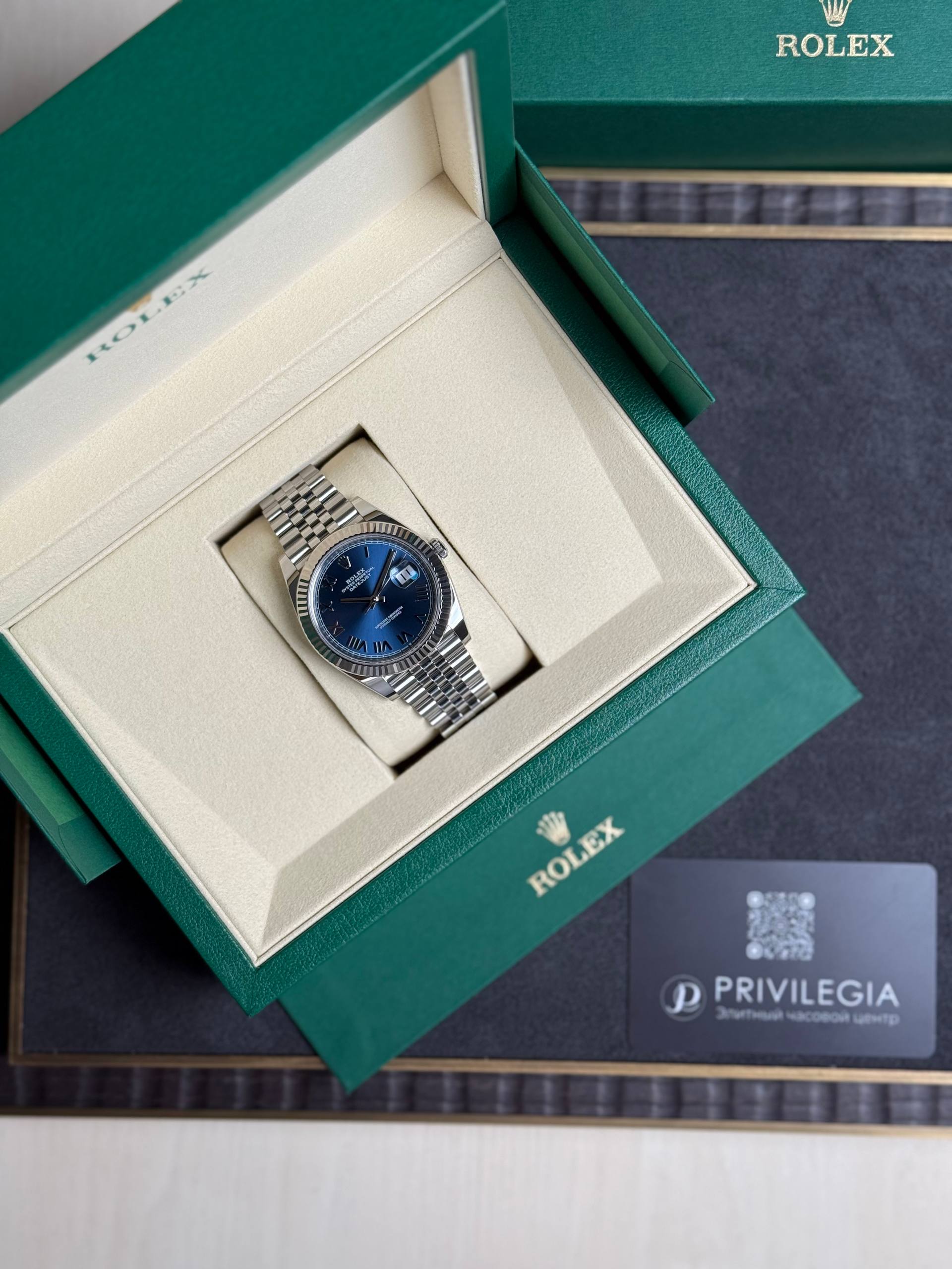 Rolex watch in an open beige interior box with a blue dial and silver bracelet, set inside a green outer box.