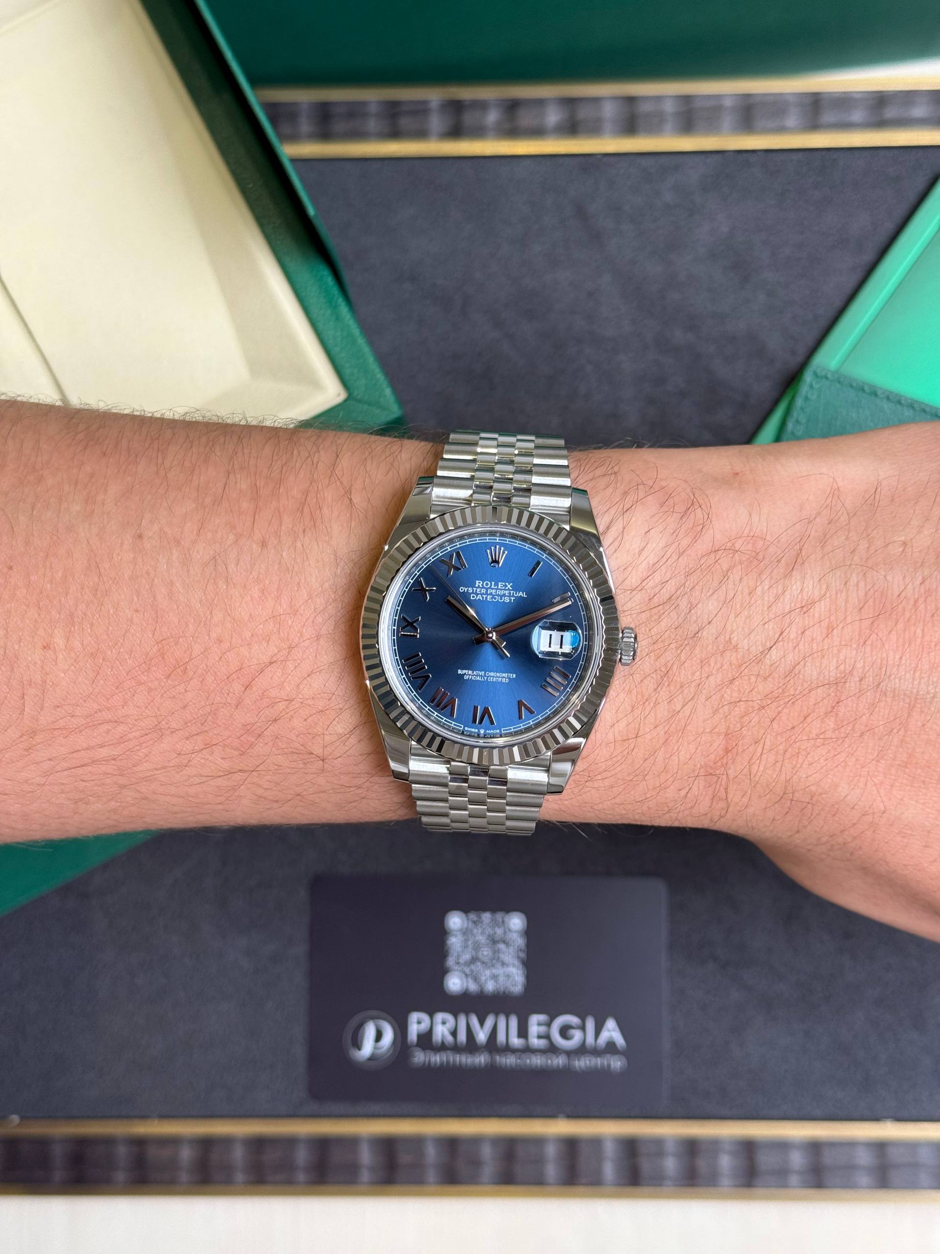 Silver Rolex wristwatch with blue dial and Jubilee bracelet on a forearm inside a green presentation box