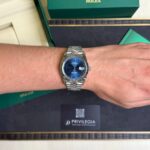 Wristwatch: stainless steel Rolex Datejust with blue dial and date window on a forearm, beside green Rolex boxes.