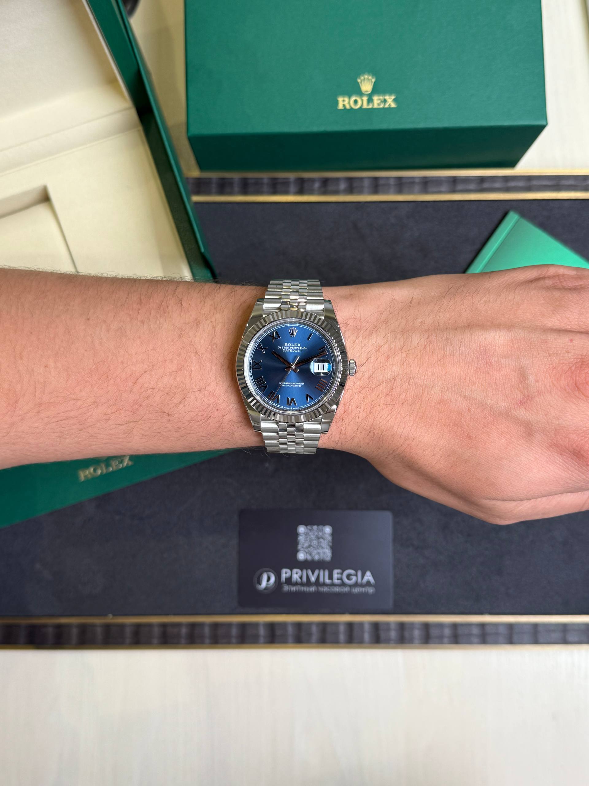 Wristwatch: stainless steel Rolex Datejust with blue dial and date window on a forearm, beside green Rolex boxes.