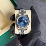 Rolex Datejust with blue dial and stainless steel Jubilee bracelet, held in a gloved hand on a beige cushion inside a green Rolex box