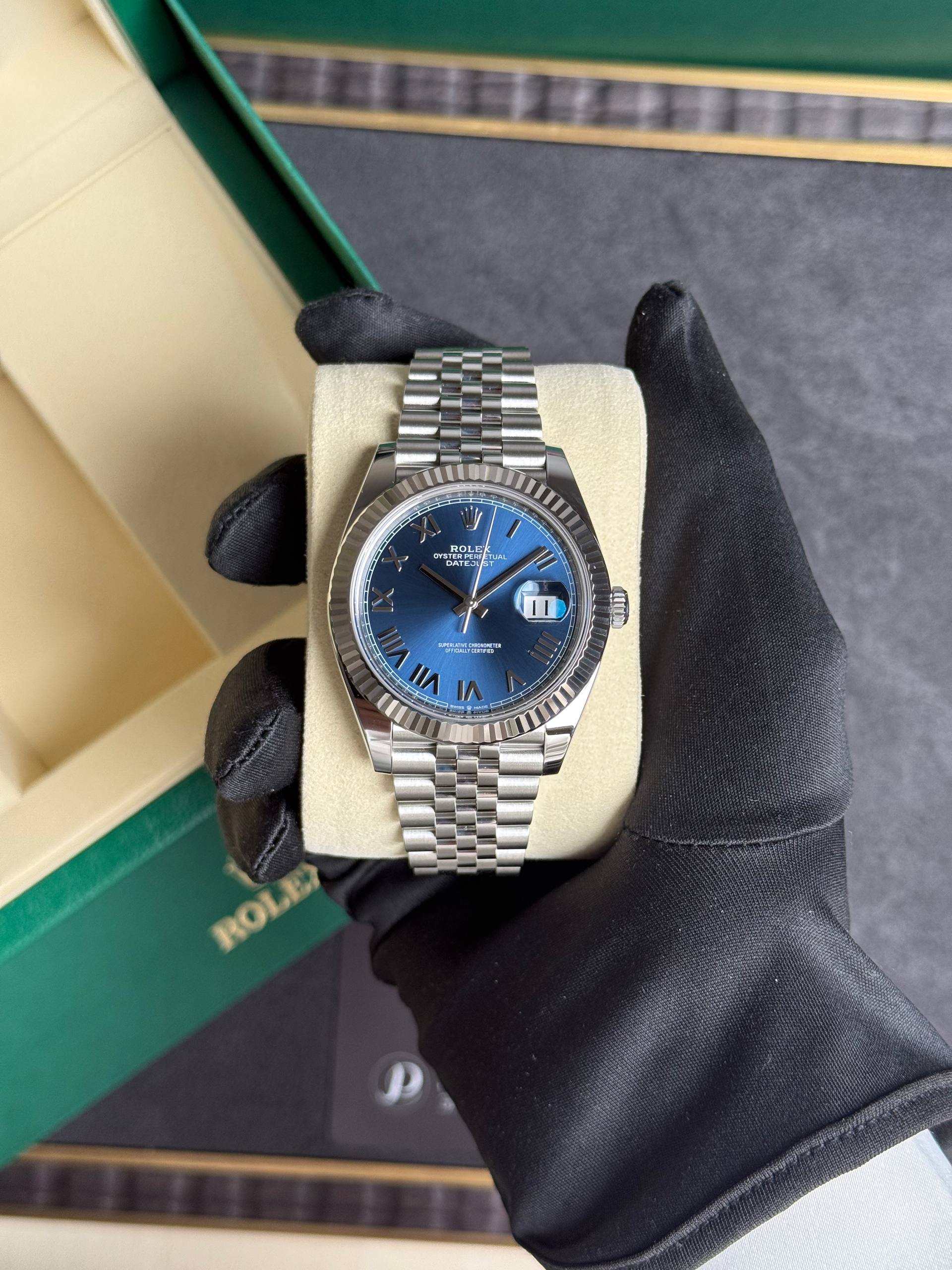 Rolex Datejust with blue dial and stainless steel Jubilee bracelet, held in a gloved hand on a beige cushion inside a green Rolex box