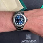 Stainless steel Rolex watch on a wrist with a blue dial and date window showing 28, next to a green box and Privilegia card.