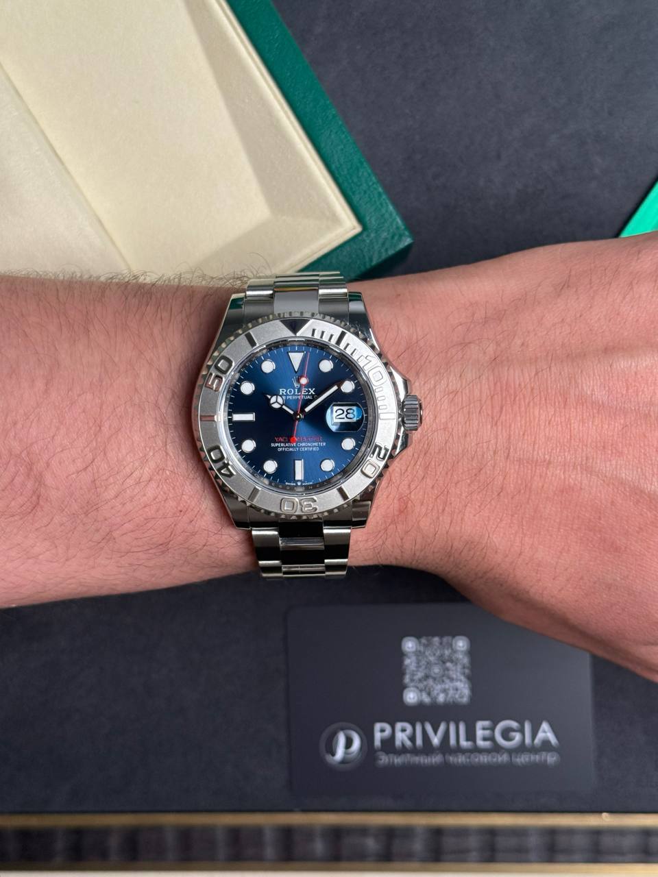 Stainless steel Rolex watch on a wrist with a blue dial and date window showing 28, next to a green box and Privilegia card.