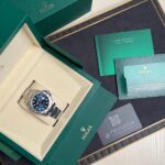 Green Rolex box opened to reveal a stainless steel watch in a beige insert, with warranty cards and booklets nearby.