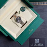 Open green Rolex box displaying a two-tone Rolex watch with black dial and date window, plus green seal tag and Privilegia card nearby.